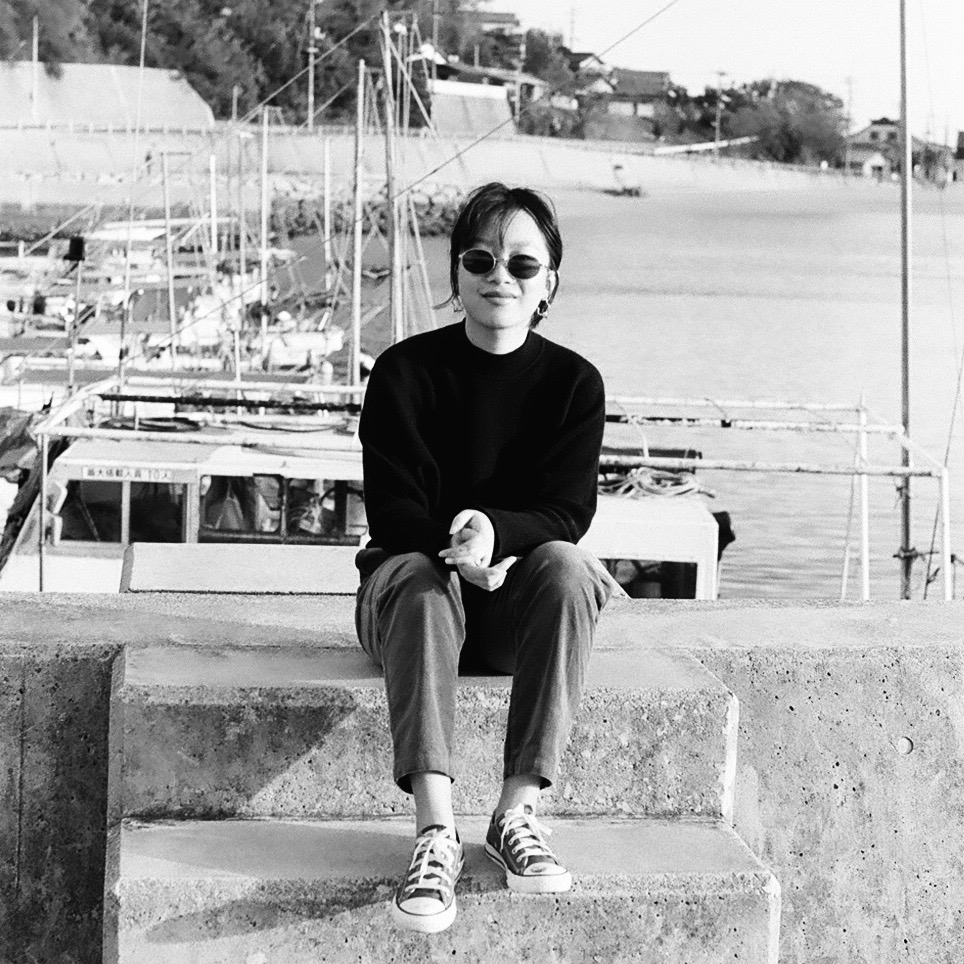Sydney Tang wears sunglasses and sits on a dock by the water.